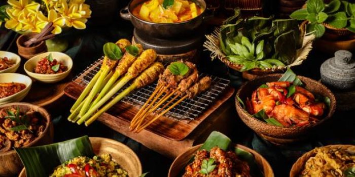 An assortment of Balinese dishes to accompany the steamed rice (not shown). The dishes are Sate Lilit (minced meat satay) and Sate Babi (pork satay), Gedang Mekuah (green papaya soup), Tum Ayam (minced chicken parcel), Udang Goreng (pan-fried prawns), Rempeyek (peanut crackers), Ayam Klungkung (chicken with crushed chili pepper), Lawar Ayam (long bean and chicken salad), Babi Goreng (seasoned pork crisps), and three chili condiments of Sambal Plecing (red chili sauce), Sambal Embe (fried red chili and shallots), Sambal Matah (shallots and lemongrass salsa). A bouquet of Frangipani is used as table decoration.